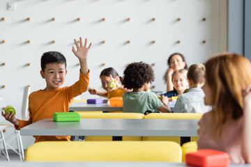 cheerful asian boy waving hand to blurred girl in school eatery