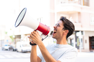 Fototapeta premium Young caucasian man at outdoors shouting through a megaphone to announce something in lateral position