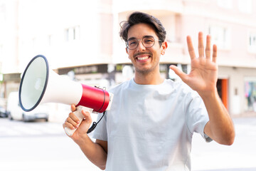Young caucasian man at outdoors holding a megaphone and saluting with hand with happy expression
