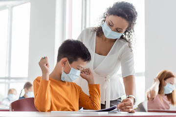 african american teacher in medical mask helping asian schoolboy during lesson