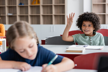 Fototapeta na wymiar smiling african american boy raising hand in classroom near blurred classmate