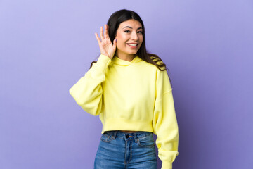 Young brunette woman over isolated purple background listening to something by putting hand on the ear