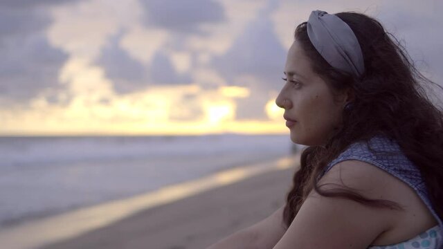 Mujer latina de perfil contempla serena y melancólica el mar al atardecer sobre la arena en la playa en Pie de la Cuesta, Acapulco, México