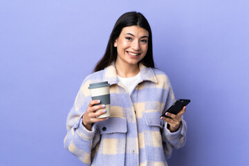 Young brunette woman over isolated purple background holding coffee to take away and a mobile