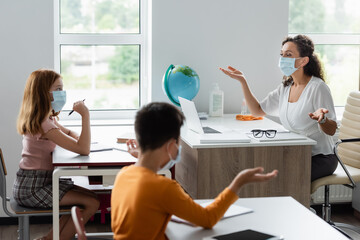 african american teacher and kids in medical masks gesturing during conversation in classroom