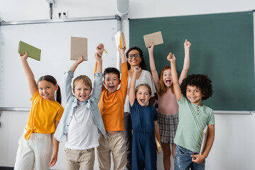 excited multiethnic schoolkids holding books in raised hands near happy african american teacher