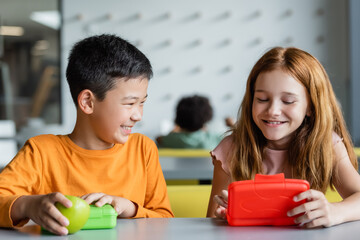 redhead girl and smiling asian boy near lunch boxes in school dining room