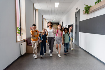 happy multicultural pupils with african american teacher walking in school corridor and waving hands
