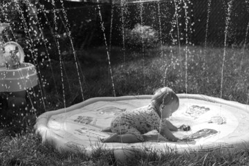 Toddler lays down on a vinyl splash pad toy in her backyard; water sprays up into the air on a hot summer day