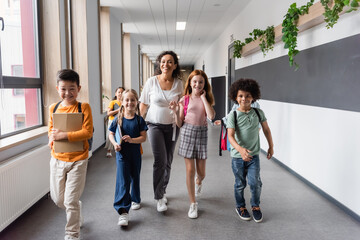 happy african american teacher walking in school corridor with multiethnic pupils