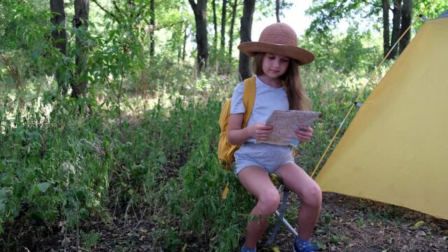 Little girl in a hat and with yellow backpack sits in the forest near the tent. Child in summer camp holding map in her hands and reading it. Concept of map reading skills for children