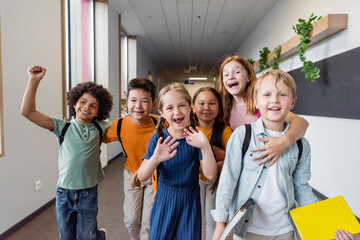 cheerful multiethnic kids embracing, waving hands and showing rejoice gesture at school
