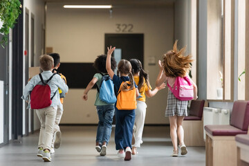 back view of multiethnic friends with backpacks running and waving hands in school corridor