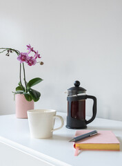 Vertical view of white cup and small coffee plunger with notebook and pen in foreground and purple moth orchid in background (selective focus)