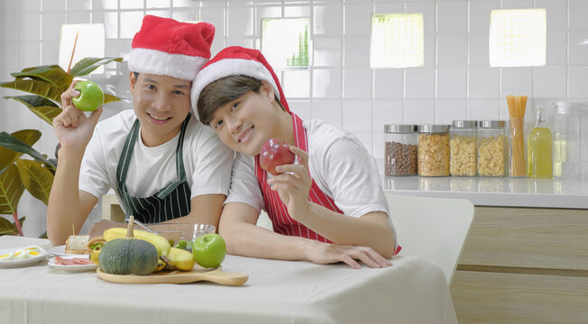 Young Asian Gay Couple Holding Apples With Smiling Face Together During Happy Meal Celebration In Dining Room On Christmas New Year Private Party At Home. Selective Focus.