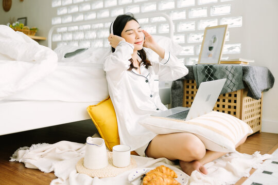 Beautiful Young Asian Woman Sitting Next To Her Bed Using Her Laptop And Enjoy Breakfast In The Morning At Home. Small Business Owners Are Checking Orders Online. Concept For Freelancer Lifestyle.