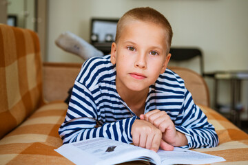 Cute ten year old boy doing homework at home on couch lying on his back during pandemic or after...
