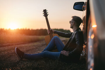 Musician with guitar sits on the dusty countryside road near the car in the sundown rays.