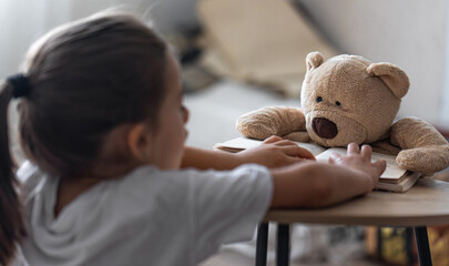 A little girl plays with her teddy bear, teaches him to read.