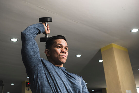 Closeup of an asian guy in a sweatshirt does single arm overhead dumbbell tricep extensions. Working out and training triceps. Open air gym setting.