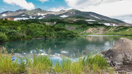 Fototapeta na wymiar Grass and wildflowers grow on the shore of the lake. A mountain range with snow on the slopes against the sky. Ripples and reflections on the water surface. A boulder in the foreground. Kamchatka
