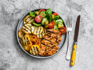 Barbecue plate - chicken breast, yellow zucchini, grilled potatoes and fresh vegetable salad with pesto sauce dressing on a grey background, top view