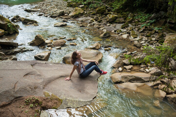 Happy smiling girl in a white t-shirt, blue jeans and red sneakers sitting near mountain river. Young woman enjoys fresh air in forest resting on a rock above a waterfall. lonely travel, side view