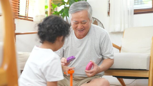 Happy Asian family having fun together at home. Smiling grandfather play toys with little grandchild girl in living room. Retired senior man teach cute baby granddaughter play colorful wooden block.