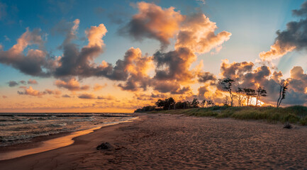 Beautiful dawn at Baltic Sea. Trees, sandy beach, water, clouds and tall grass illuminated by...