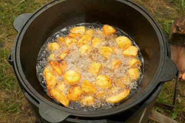 Potato is frying in cauldron in oil on nature on fire, closeup view. Preparing food on campfire in camping. Large pieces of potatoes are deep-fried in a black kazan. Cooking using wood-burning stove.