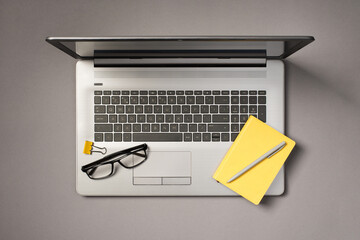 Top view photo of glasses yellow binder clip pen and yellow notepad on open grey laptop on isolated grey background