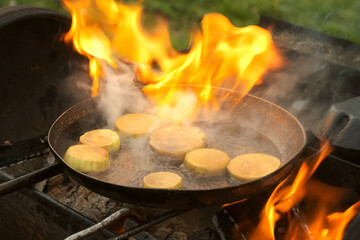 Man is frying slices of fresh organic zucchini on frying pan with hot boiling oil on coals in grill. He is putting zucchini in hot oil and its starting to burn. Preparing food in chargrill on nature.