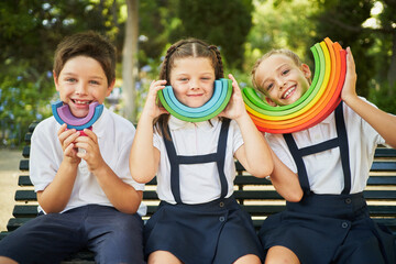 Fototapeta na wymiar Three children wearing school uniform smiling at camera with rainbow toy in park