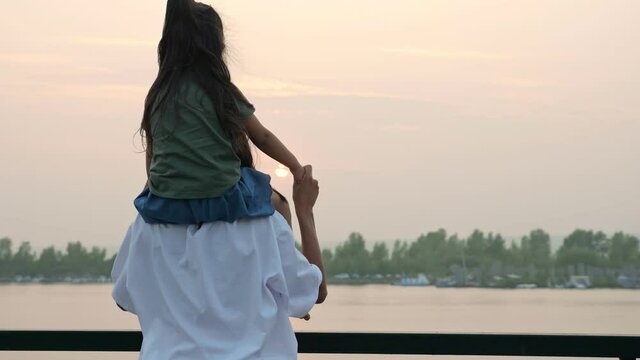 Mommy in loose t-shirt holds toddler daughter on shoulders looking at calm river on city embankment in summer evening twilights backside view