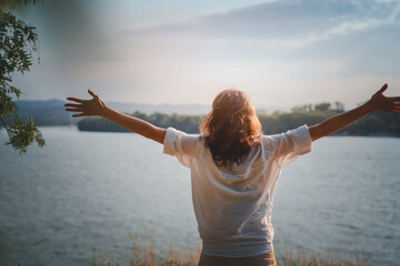 Happy beautiful young woman enjoying the view and landscape on the river bank with hands raised up at sunset