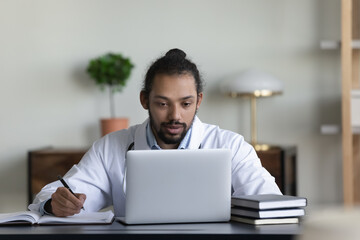 Concentrated young african american mixed race general practitioner doctor working on computer in modern clinic office, handwriting notes in journal, managing appointments or consulting distantly.