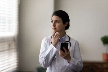 Thoughtful young indian female general practitioner doctor holding cellphone in hands looking in...