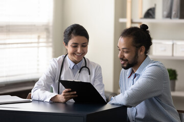 Happy young indian female gp doctor showing test result on clipboard to interested millennial african ethnicity male patient, discussing healthcare medical insurance or illness treatment at clinic. - Powered by Adobe