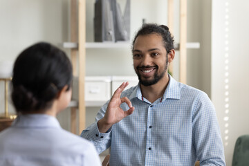 Smiling young handsome african american man using sign language, practicing communication with professional female indian ethnicity therapist, showing symbols with fingers in clinic meeting.