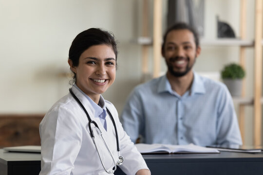 Portrait Of Smiling Confident Young Female Indian Ethnicity Gp Doctor Physician Posing In Clinic With Blurred African American Male Patient On Background, Professional Medicare Healthcare Services.