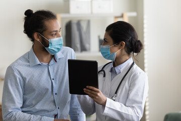 Focused young mixed race diverse male patient and female gp doctor discussing illness treatment, healthcare medical insurance benefits or regular checkup tests results, holding digital computer tablet - Powered by Adobe