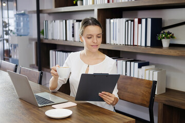 Smiling Caucasian woman happy with a cup of hot coffee in the morning at cafe. She is relaxing with online community on laptop and work sheet in cafe. Lifestyle of businesswoman and technology concept