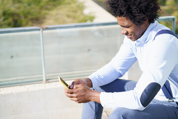 Black man with afro hairstyle using a smartphone sitting near an office building.