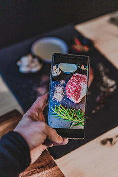 People is taking a photo fresh raw rib eye steak on wooden cutting Board  with salt, pepper, olive oil,garlic and rosemary.