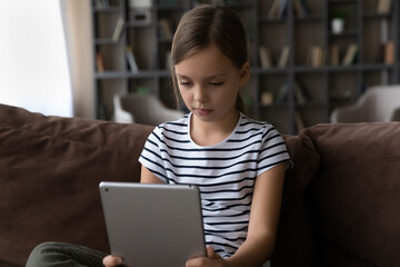 Adorable small preteen child girl using digital computer tablet, involved in laying games or web surfing photo video content in social network, modern tech addiction, internet security concept.