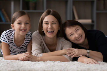 Fototapeta premium Portrait of happy adorable small kid girl lying on floor carpet with affectionate loving mother and laughing older mature grandmother, enjoying spending carefree weekend leisure time together at home.