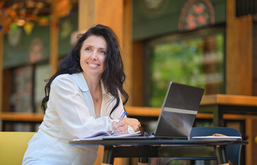 Mature woman at coffee shop. Portrait of confident mature professional woman sitting on summer terrace in cafe, using laptop computer for work, laughing happily indoors.