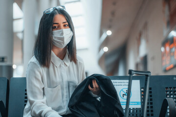 Chica joven sonriente con mascarilla y equipaje en el aeropuerto cumpliendo el distanciamiento...
