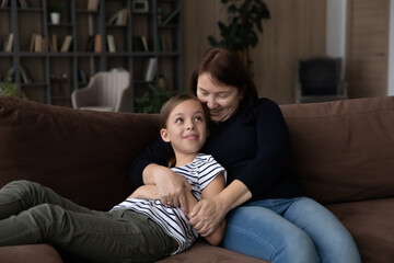 Happy caring middle aged older granny embracing smiling small cute grandchild, enjoying communicating relaxing together on comfortable couch, talking speaking chatting together on weekend at home.