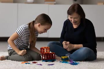 Cheerful small preteen kid girl playing magnetic plastic constructor with caring middle aged senior retired grandmother, sitting on comfortable floor carpet in modern living room, family playtime.